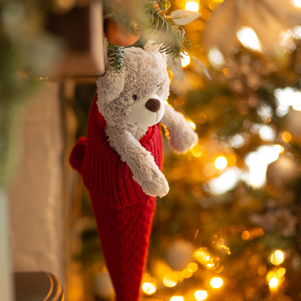 A soft toy bear tucked inside a red knitted stocking hanging by a Christmas tree with warm golden lights in the background.