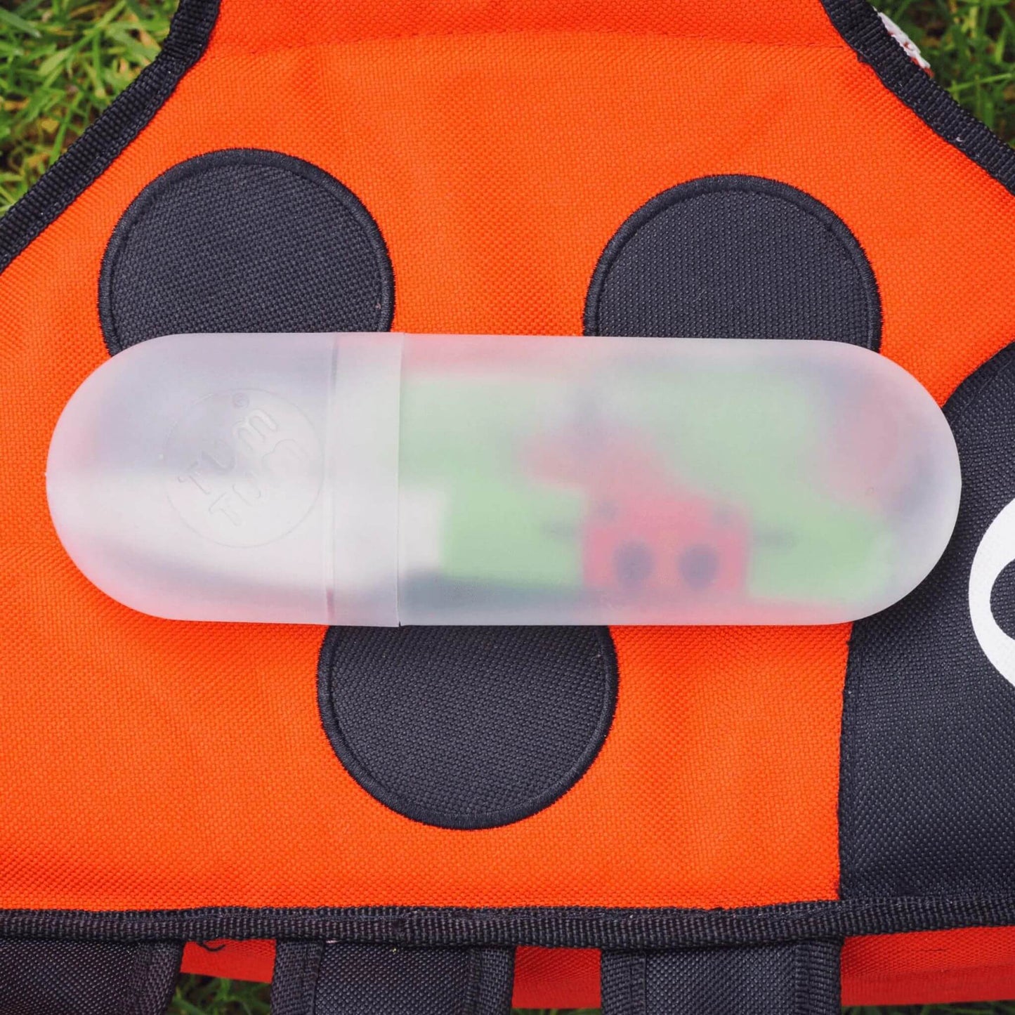 A toddler's cutlery set stored in its frosted travel case, placed on a red and black ladybird themed backpack.