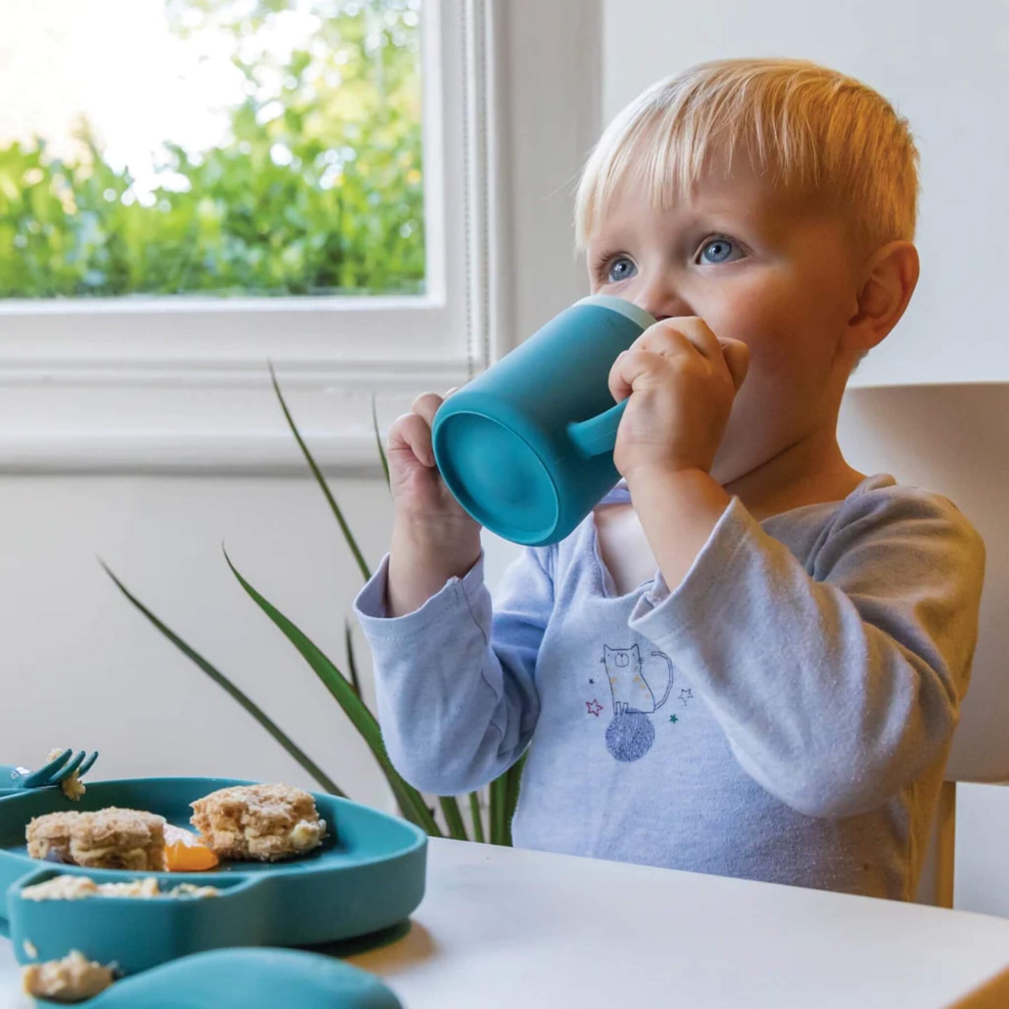 A toddler drinking from a blue silicone open cup with two handles while seated at a table during mealtime.