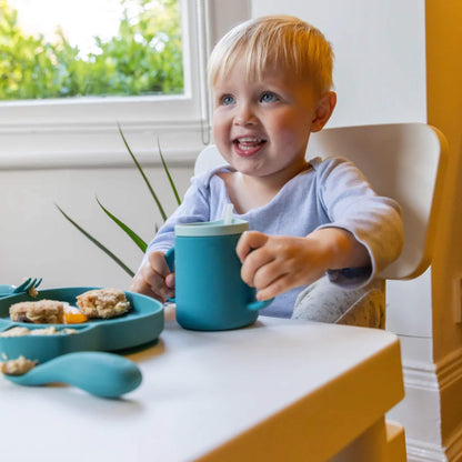 A toddler seated at a highchair holding a blue silicone trainer cup with two handles, during a meal.