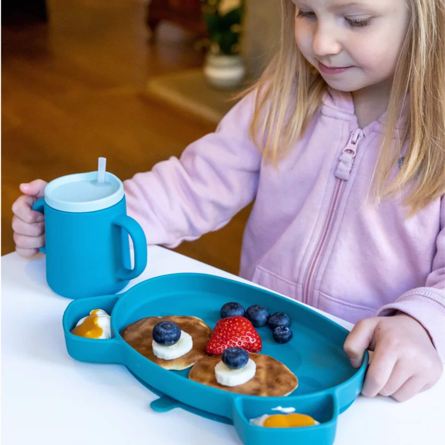 A young child holding a blue silicone trainer cup with a straw lid beside a matching silicone plate during a meal.