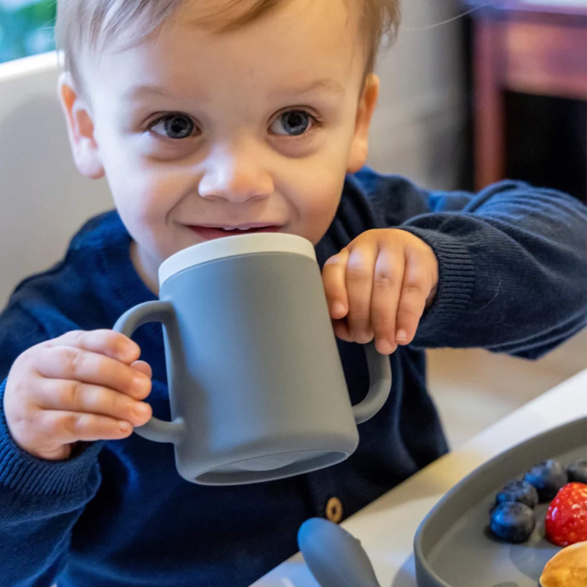 A toddler seated at a table holding a grey silicone cup with two handles, bringing the rim to their mouth while looking forward.