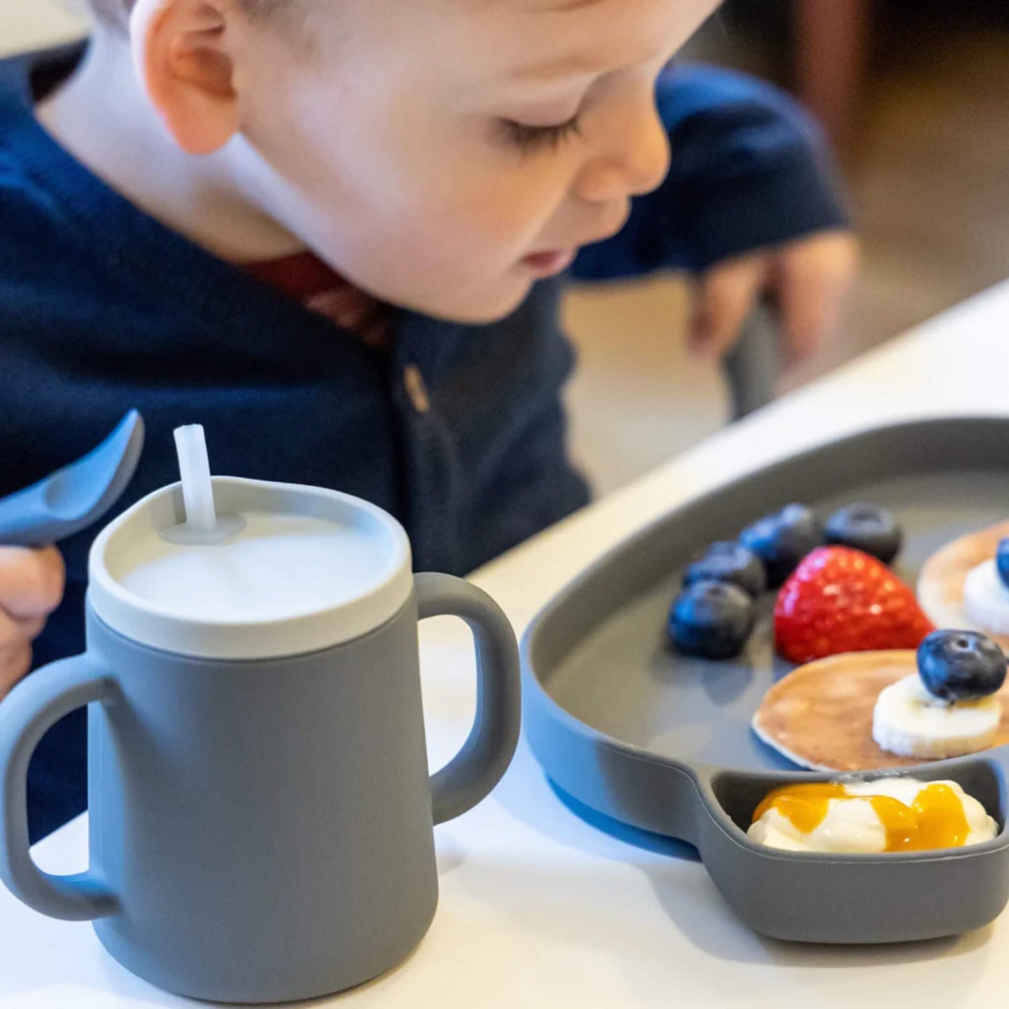 A toddler seated at a table with a grey silicone cup placed beside a sectioned plate containing fruit and small pancakes, viewed from the side.