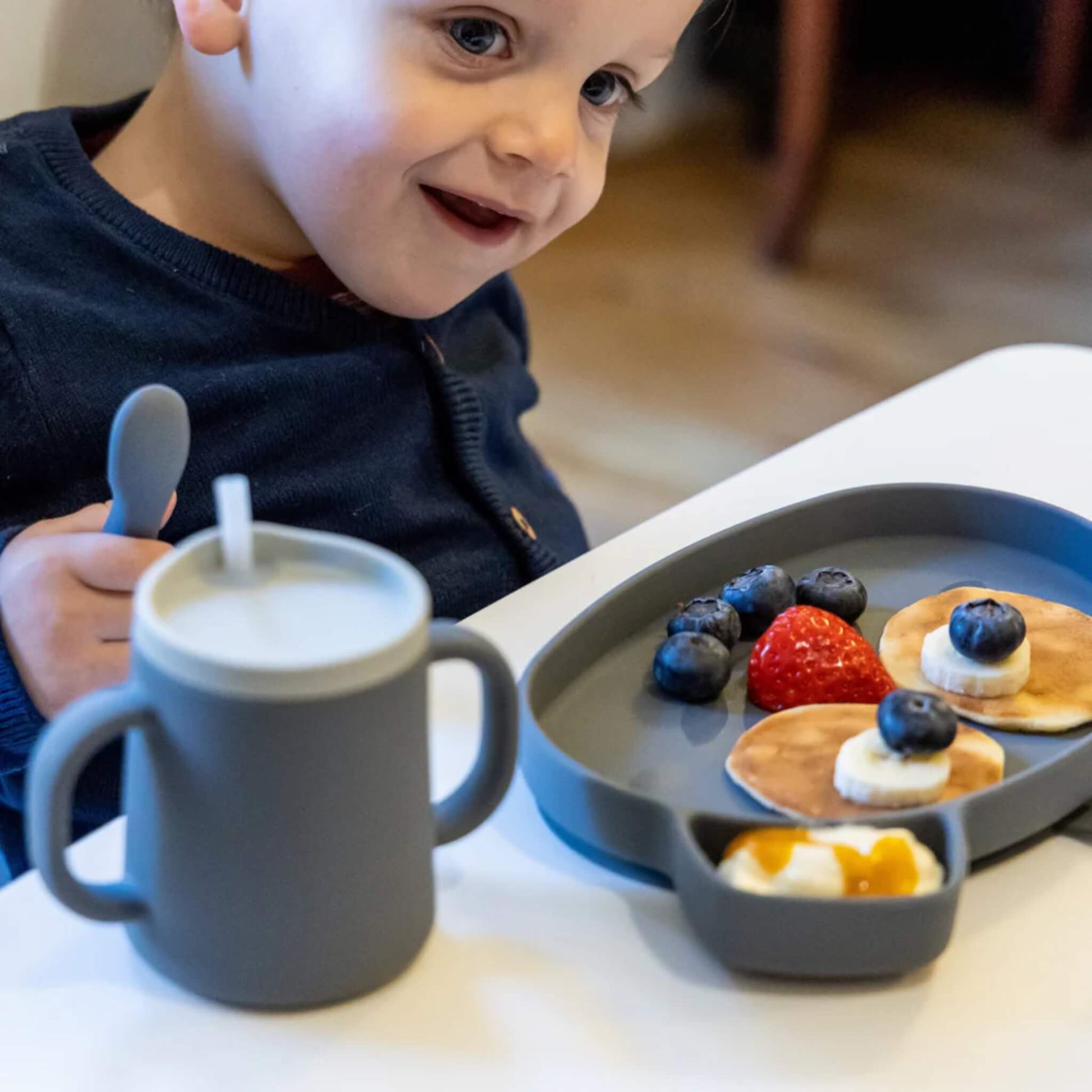 A young child seated at a table with a grey silicone cup fitted with a straw lid placed in front, alongside a grey divided plate holding pancakes, fruit and yoghurt.