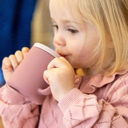 Young child drinking from a pink silicone trainer cup with two handles, holding the cup close to their mouth.