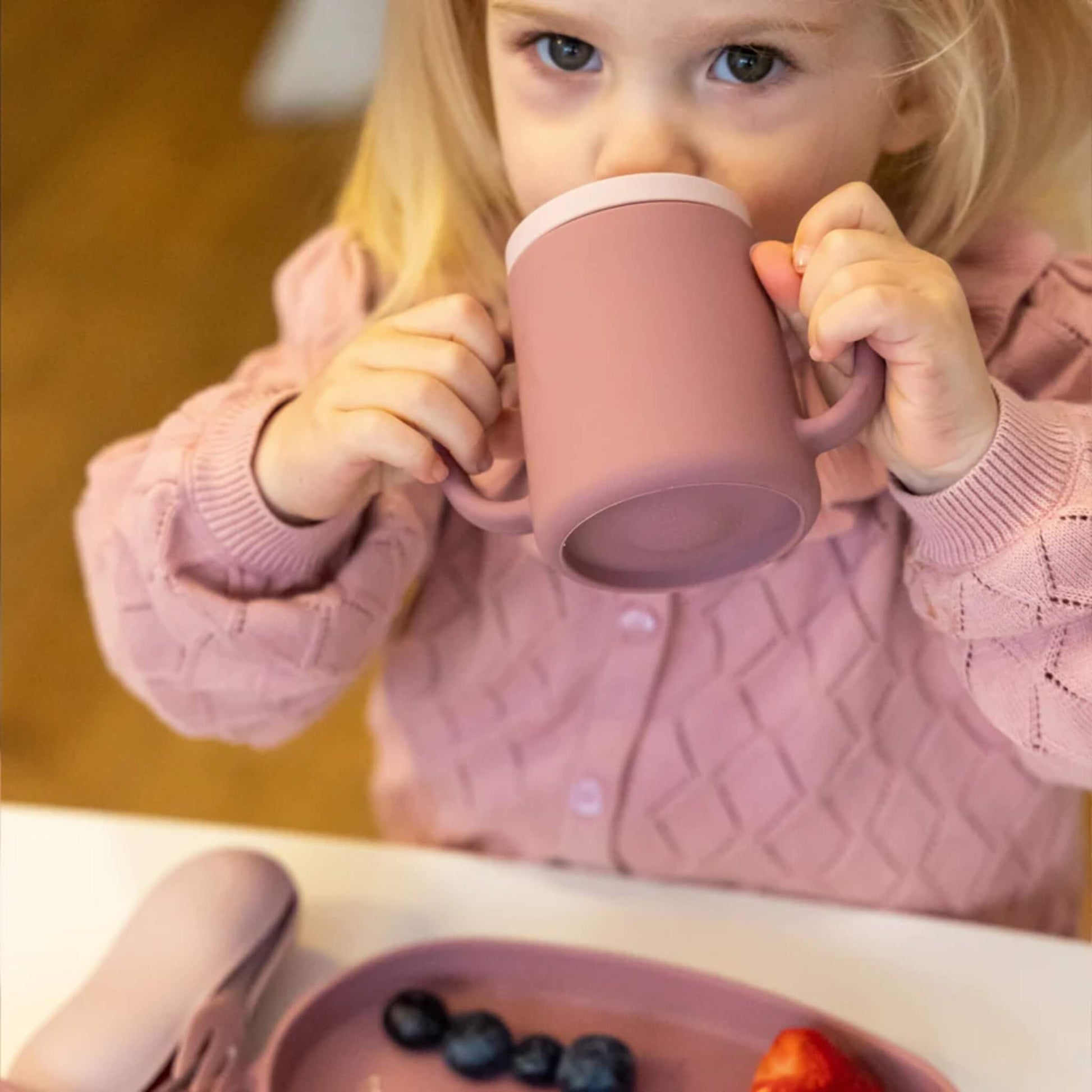 Front view of a young child drinking from a pink silicone trainer cup with two handles, with the cup tilted towards their mouth.