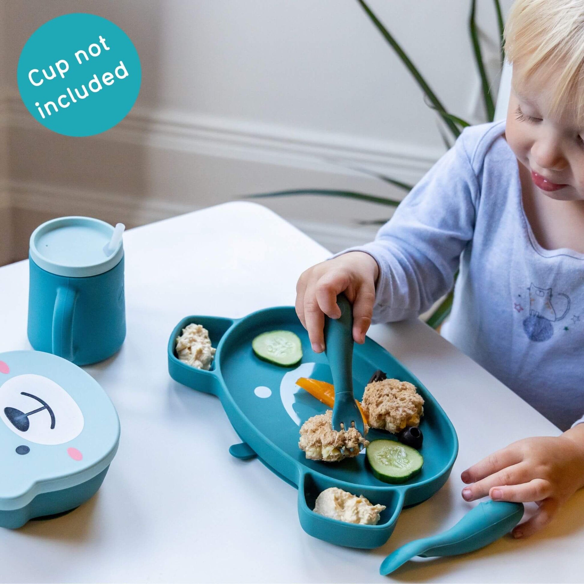 Toddler using a bear-shaped plate and cutlery set to eat sandwiches and vegetables, with a matching snack pot and cup.