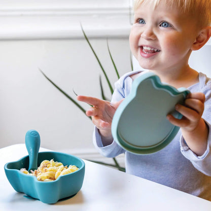 Toddler smiling while holding a blue bear-shaped lid. A silicone bowl filled with pasta sits on the table alongside a matching spoon.