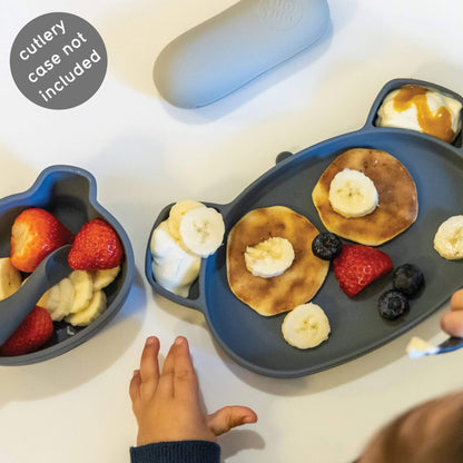 Toddler using a grey bear-shaped plate and snack pot with pancakes, fruit, and yoghurt. Designed for self-feeding.
