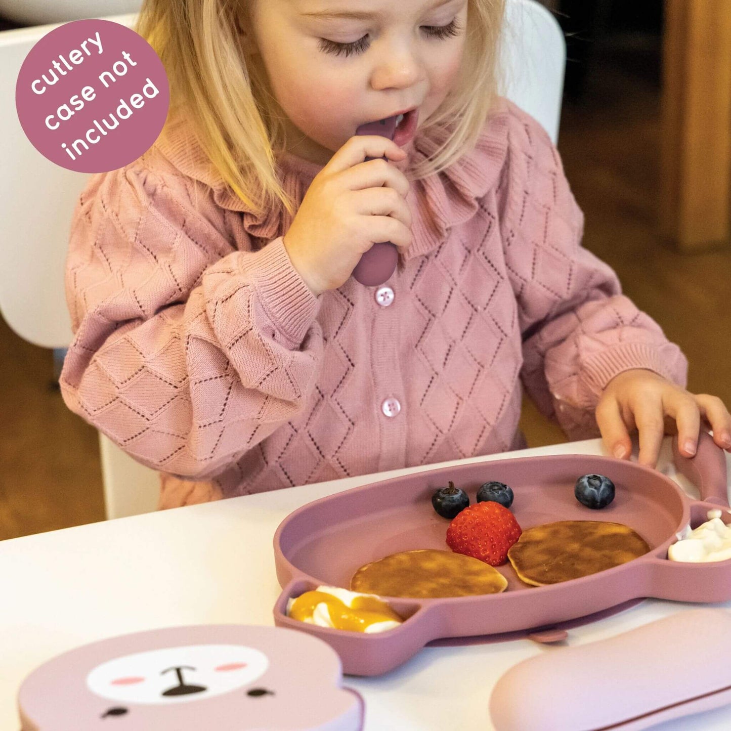 Toddler using pink silicone cutlery and plate with pancakes, fruit, and yoghurt, wearing a pink jumper, perfect for self-feeding.