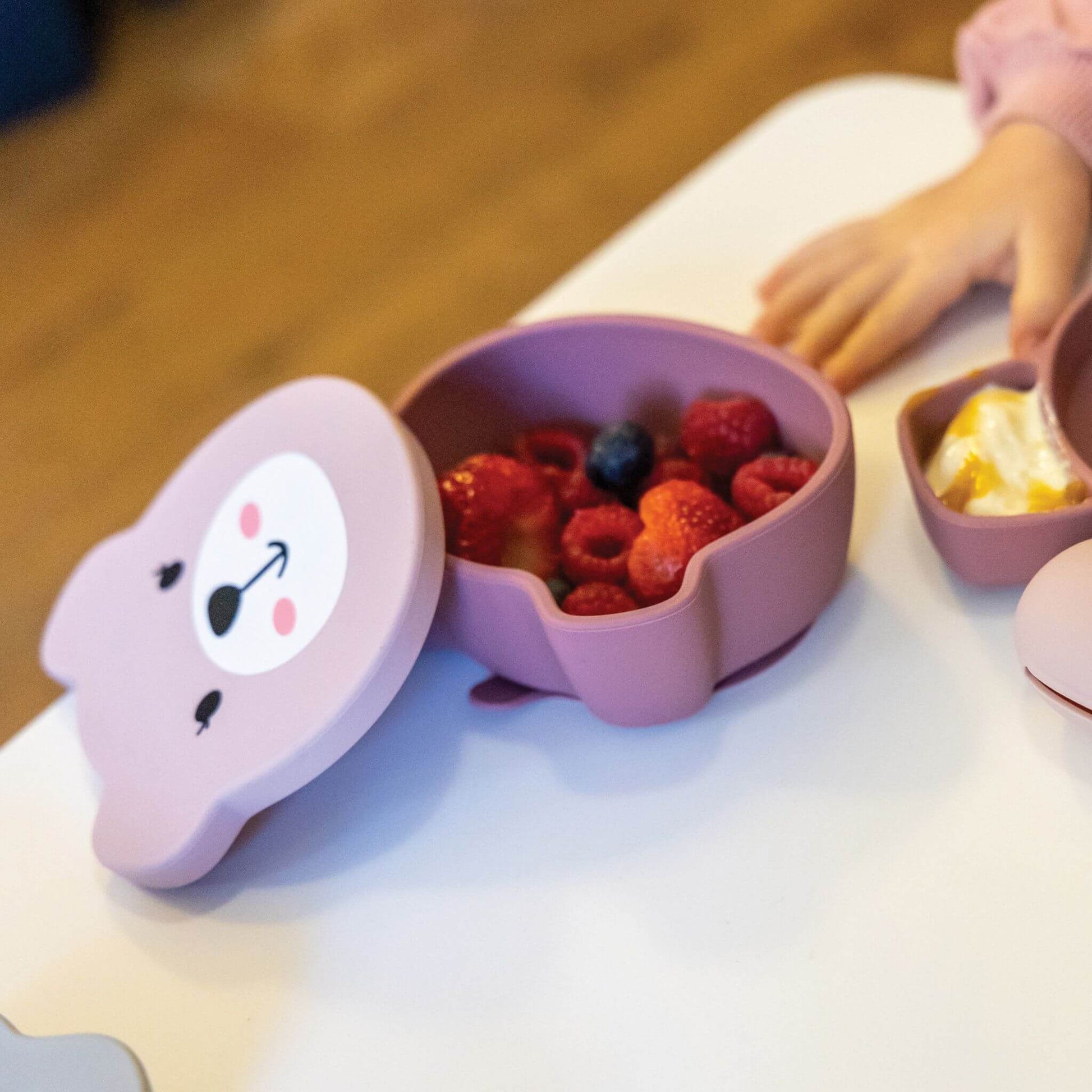 Pink silicone bear-shaped snack pot filled with fresh berries, alongside a matching plate with yoghurt, perfect for toddlers learning to self-feed.