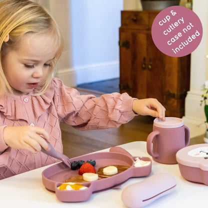 Toddler using pink silicone cutlery, plate, and cup with pancakes, fruit, and yoghurt, wearing a pink jumper, ideal for self-feeding