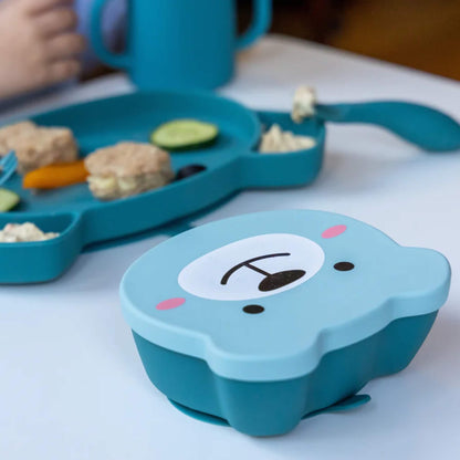 Blue silicone bear-shaped snack bowl placed on table beside a matching blue divided suction plate with finger foods.