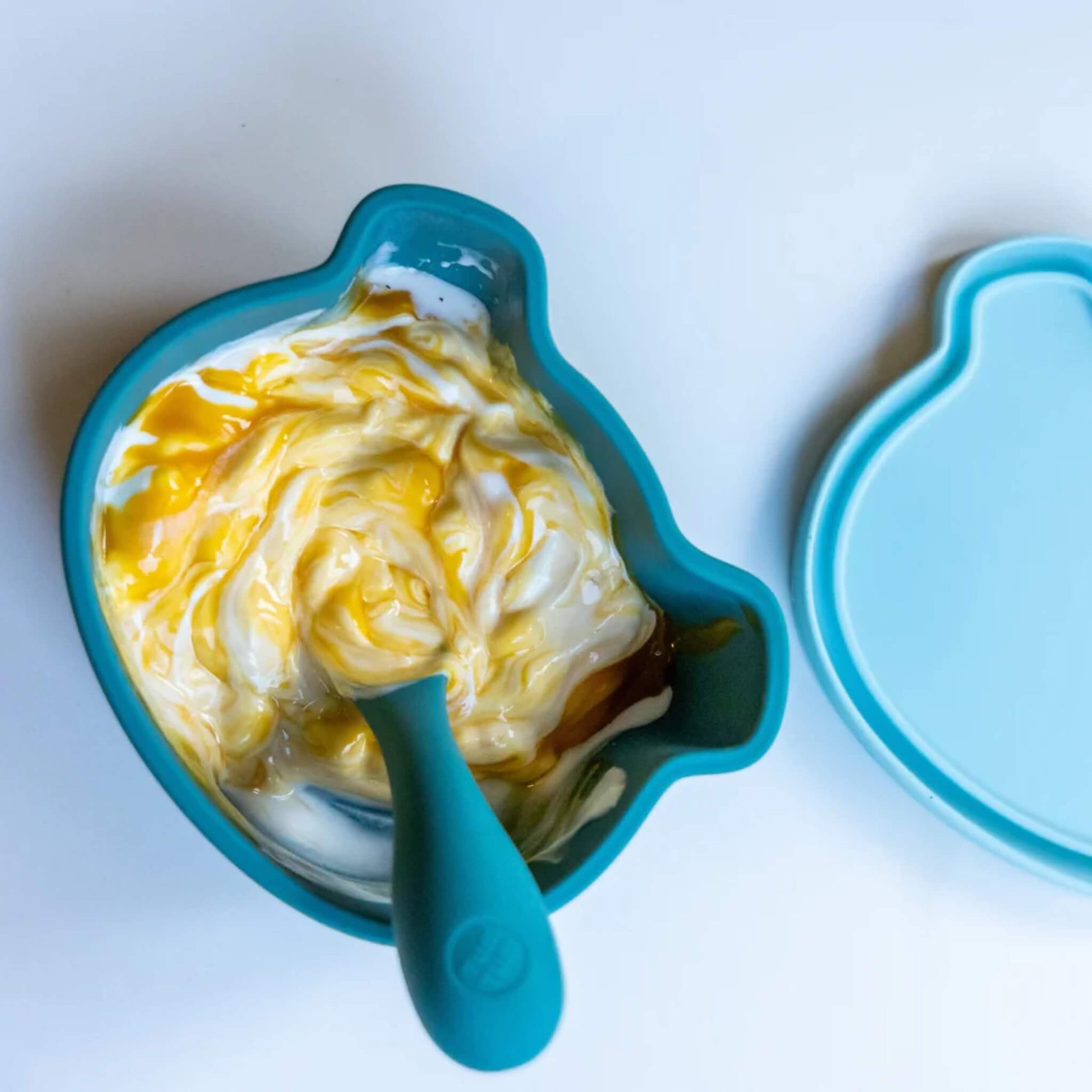 Blue silicone baby bowl filled with yoghurt and fruit purée, with chunky handled spoon resting inside bowl.