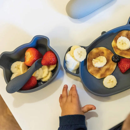 A baby's hand reaching towards a sectioned silicone plate and bowl set on a table, with soft finger foods arranged for self-feeding.