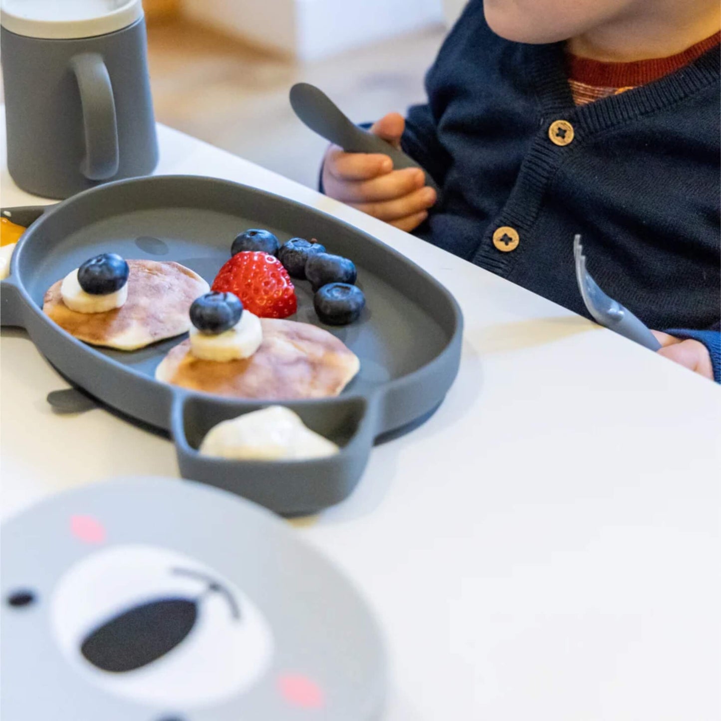 A baby holding a chunky silicone spoon while seated at a table with a suction plate holding fruit and soft foods.