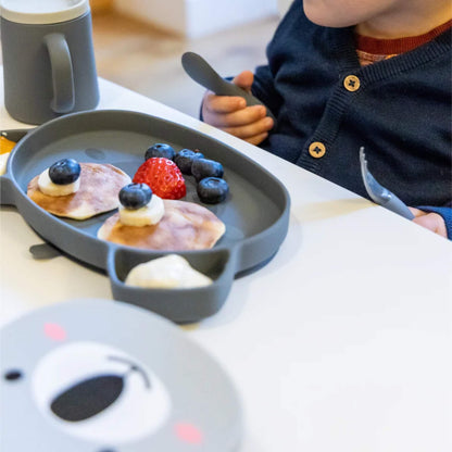 A baby holding a chunky silicone spoon while seated at a table with a suction plate holding fruit and soft foods.