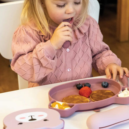 A toddler seated at a table using a chunky handled spoon with a sectioned silicone plate holding pancakes, fruit and yoghurt.
