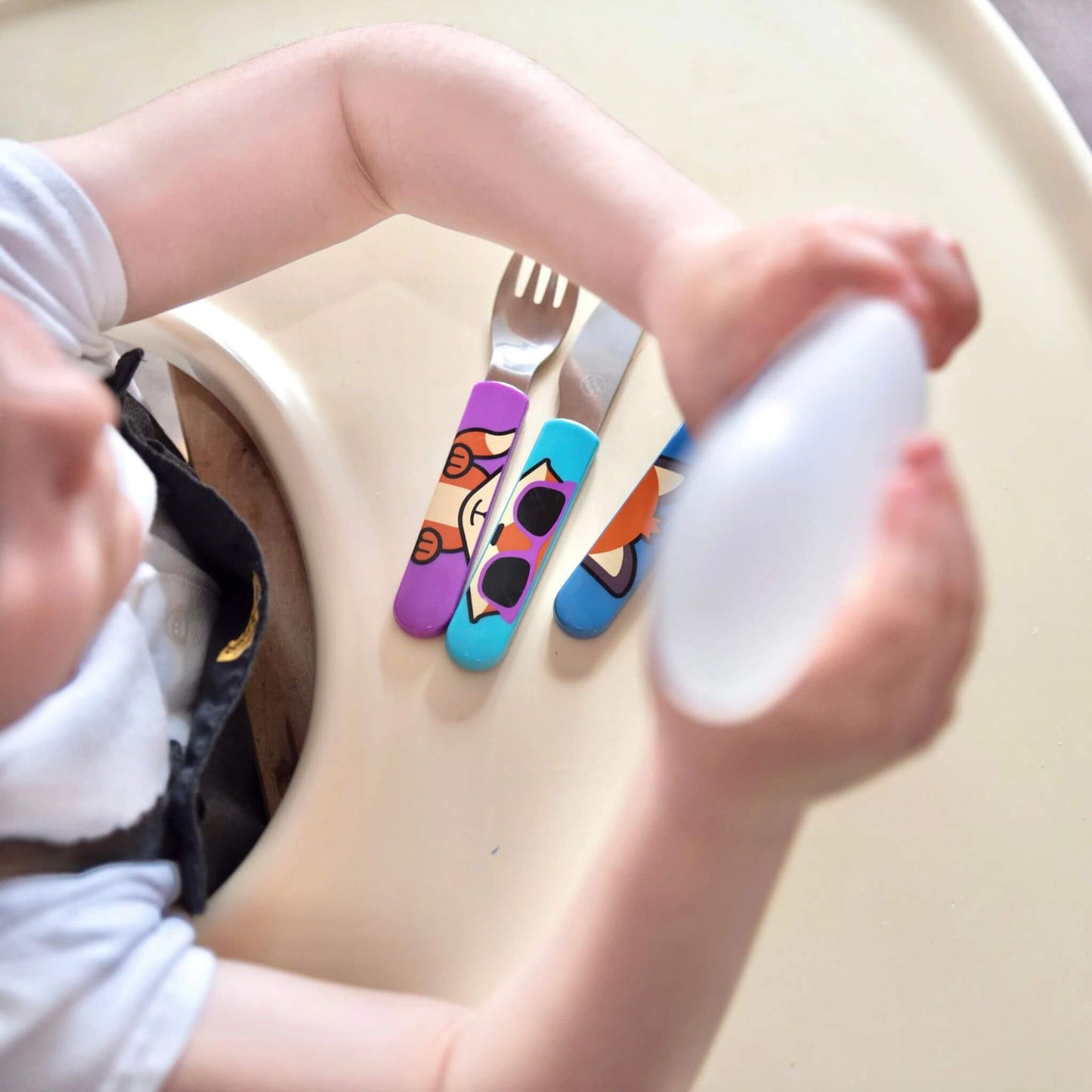 A child seated at a high chair with a cutlery set, featuring colourful Felicity Fox handles on the fork, knife, and spoon. Designed for easy self-feeding.