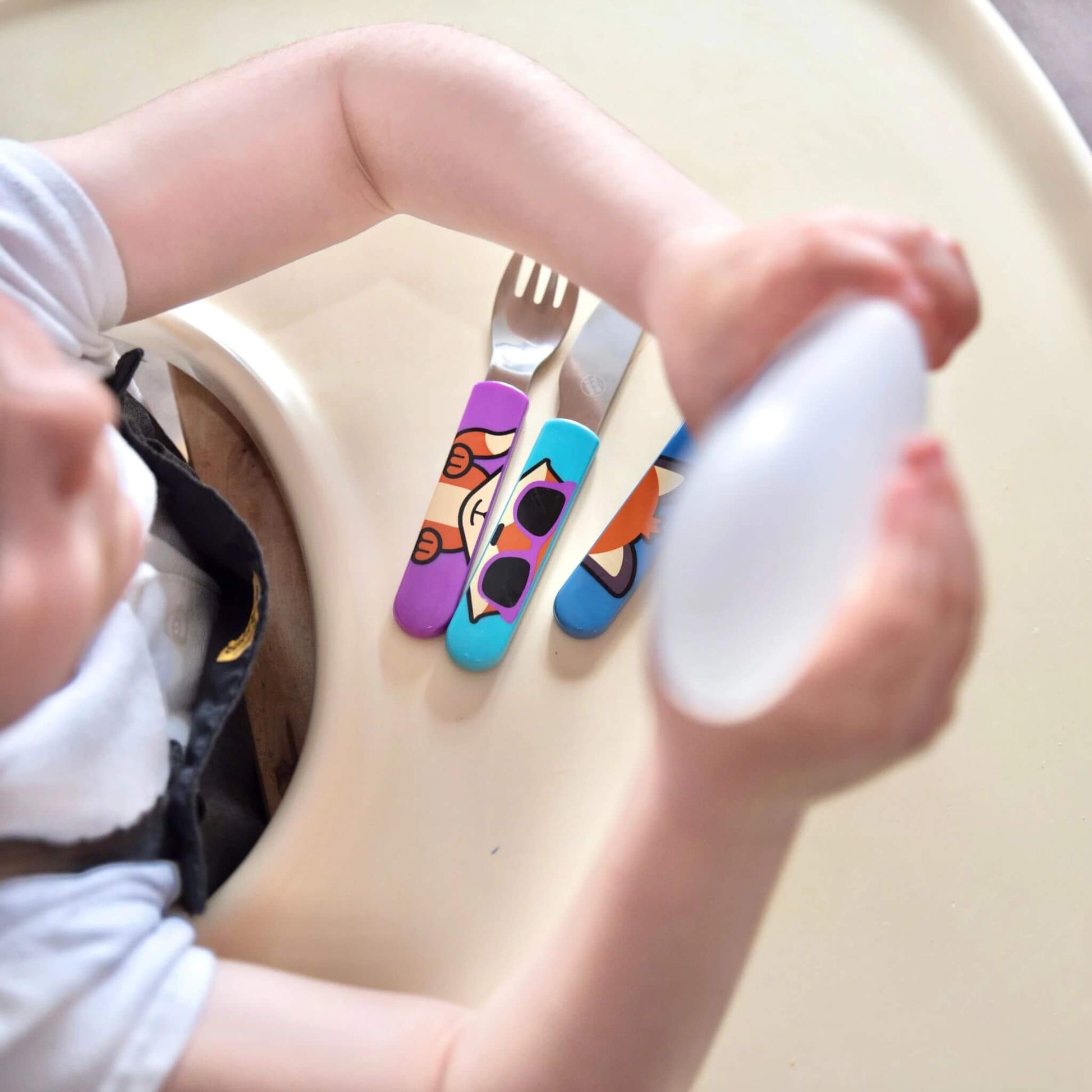 A child seated at a high chair with a cutlery set, featuring colourful Felicity Fox handles on the fork, knife, and spoon. Designed for easy self-feeding.