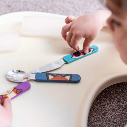 A child playing with a cutlery set, featuring Felicity Fox handles on the spoon, fork, and knife. Designed for easy grip and self-feeding.
