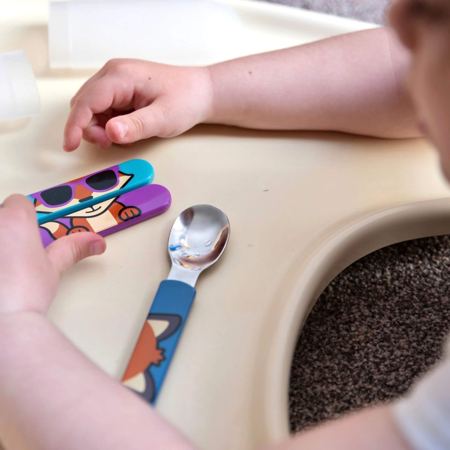 A child interacting with cutlery set, which includes a spoon, knife, and fork with vibrant Felicity Fox handles. Designed for a comfortable grip and to encourage self-feeding.