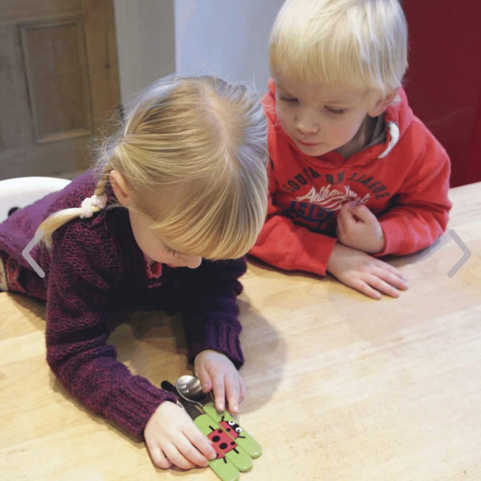 Two children examining a kids cutlery Set, featuring bright green handles with a ladybird design. Ideal for encouraging self-feeding.