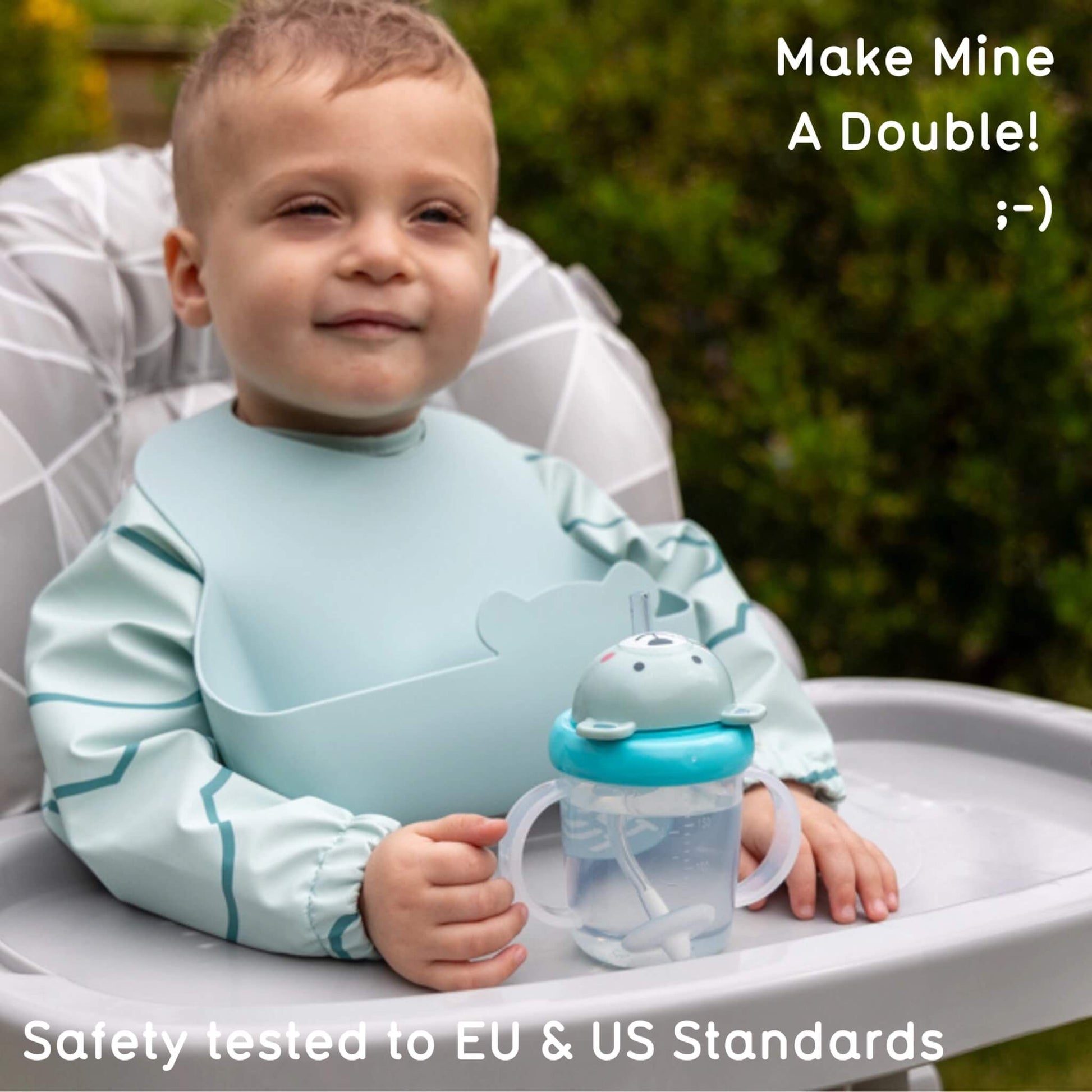 A baby sitting in a high chair outdoors holding a sippy cup, wearing a light blue bib. Text overlays read 'Make Mine A Double! ;)' and 'Safety tested to EU & US Standards'.