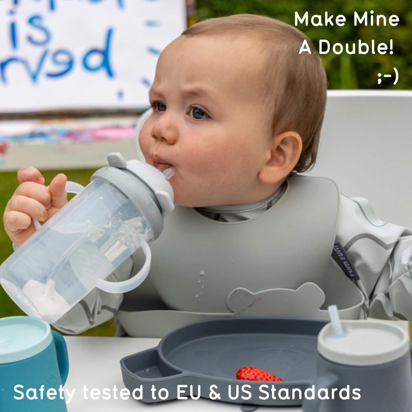A baby wearing two grey bibs drinks from a handled straw cup at an outdoor table, with grey and blue children’s tableware arranged in front.
