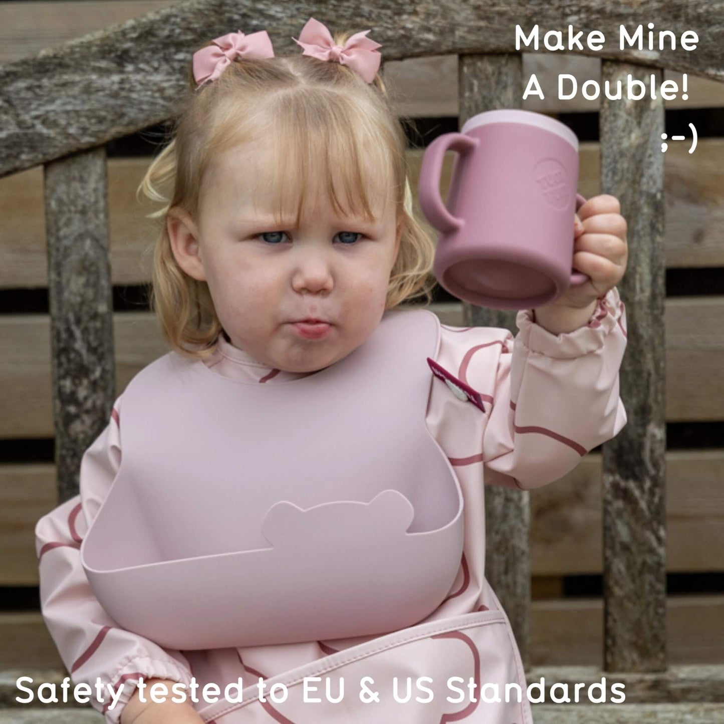 A young child sitting on a wooden bench holding a dusty pink handled cup, wearing a matching dusty pink bib and pink outfit with bow hair accessories. Text overlays read 'Make Mine A Double!' and 'Safely tested to EU & US Standards'.