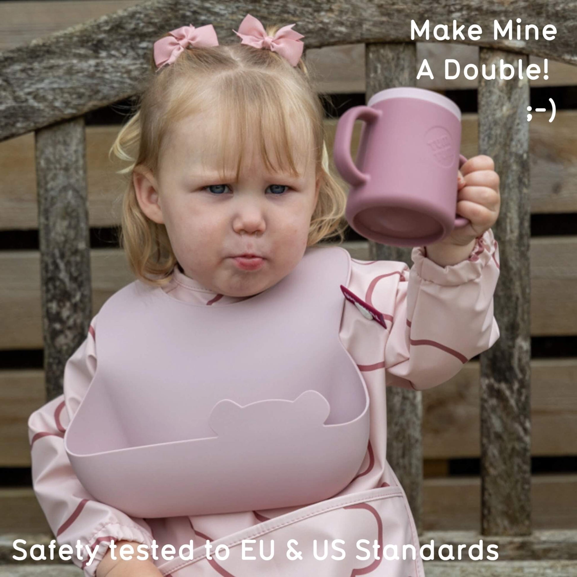 A young child sitting on a wooden bench holding a dusty pink handled cup, wearing a matching dusty pink bib and pink outfit with bow hair accessories. Text overlays read 'Make Mine A Double!' and 'Safely tested to EU & US Standards'.