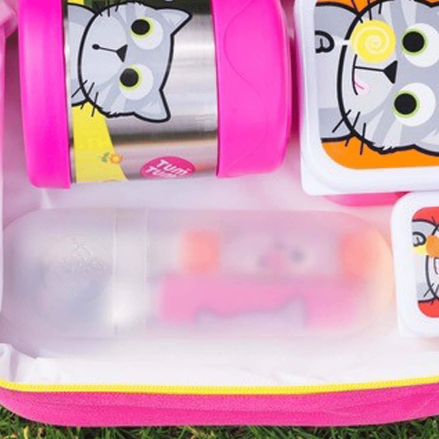 A pink and white lunch bag containing a stainless steel thermos with a cartoon cat design, a clear plastic bottle, and matching snack containers, photographed outdoors on grass.