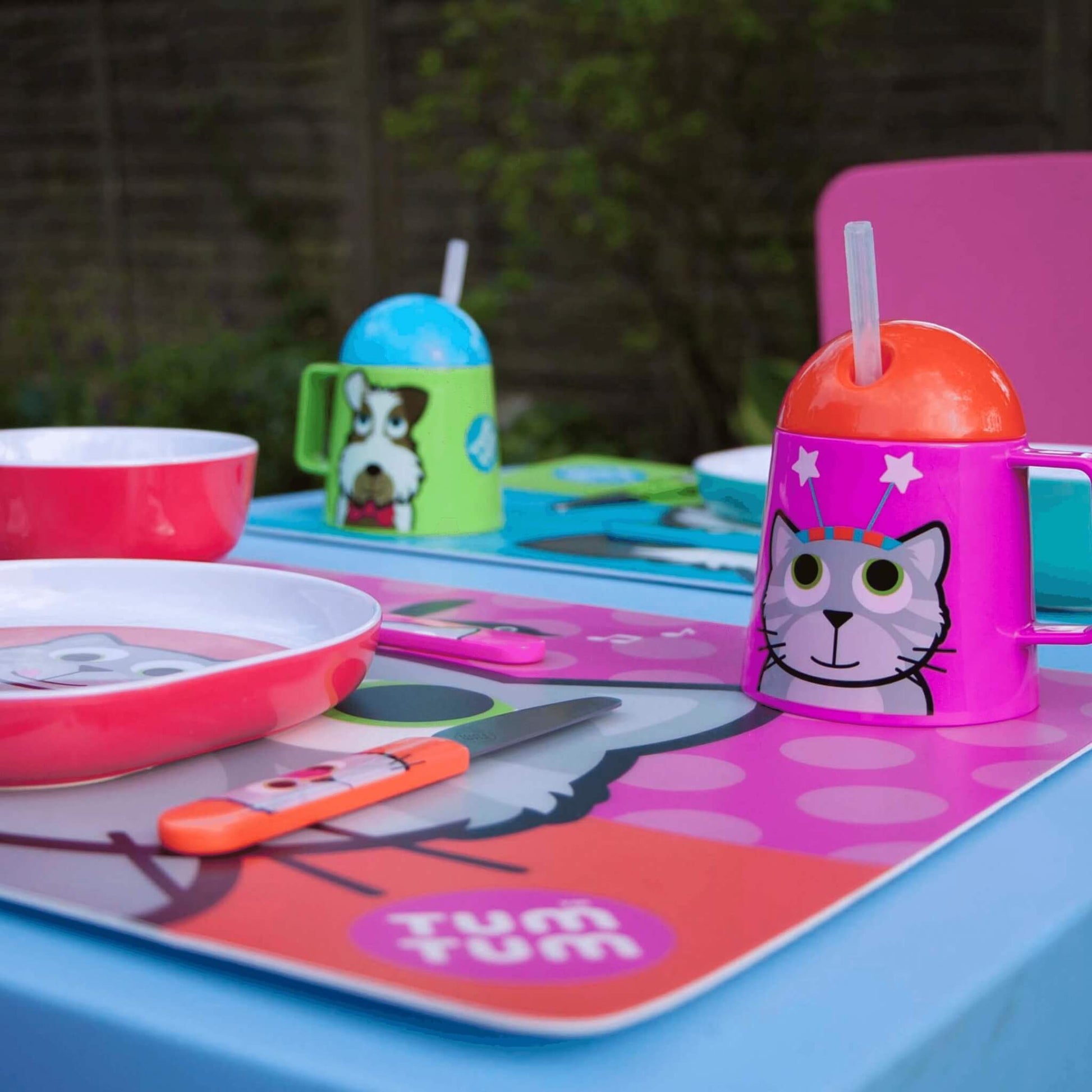 Children's tableware set with colorful cups, plates, and cutlery on a pink mat with 'Tum Tum' branding.