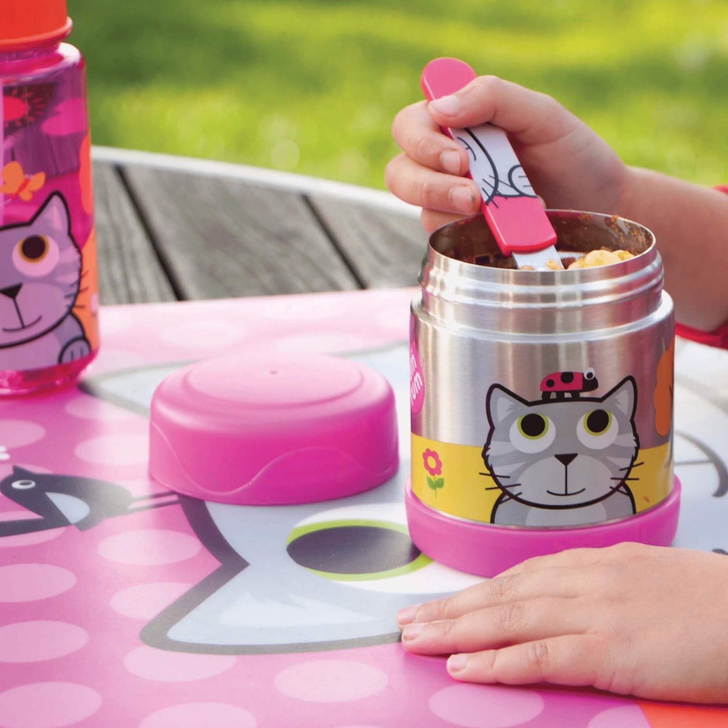 A stainless steel lunch container with a pink lid and a cat illustration, shown outdoors on a pink table mat with a child's hands interacting with it.