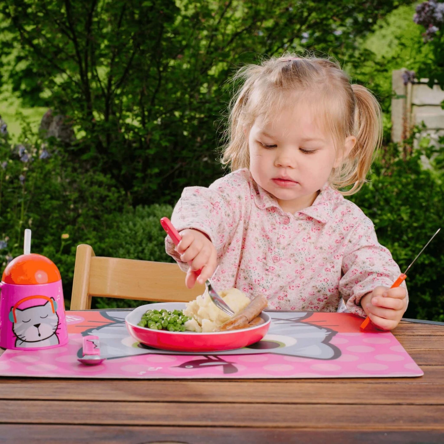 A young toddler girl with blonde pigtails sits at an outdoor wooden table, eating mashed potatoes and peas from a pink plate. She is wearing a pink floral long-sleeve shirt and holding a pink-handled fork and a knife.