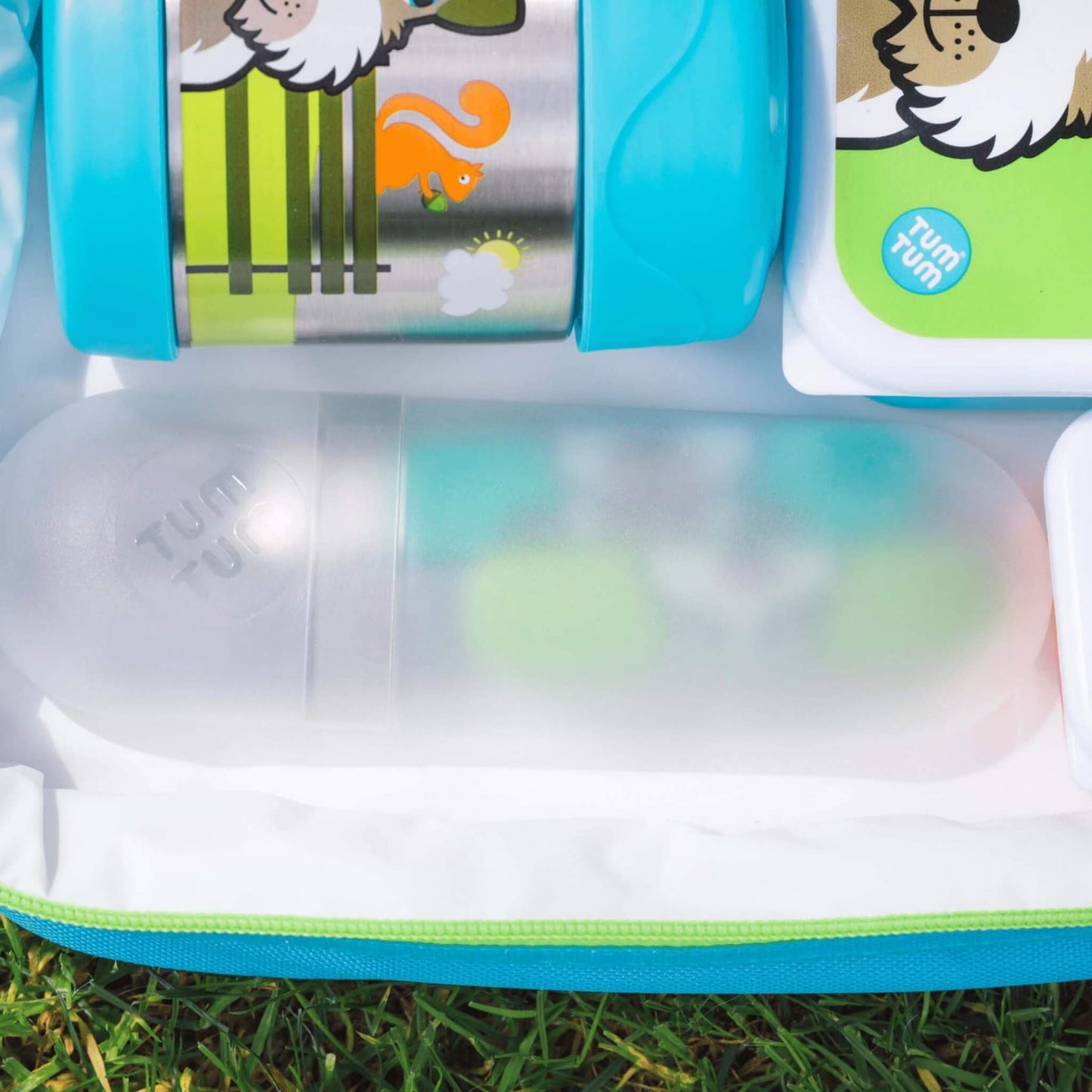 A close-up of a Tumbler insulated lunch bag or organizer on grass, showing a clear plastic bottle with a translucent cap, colorful cartoon lunch containers with bright blue lids, and a white pouch with a green Tumbler logo.