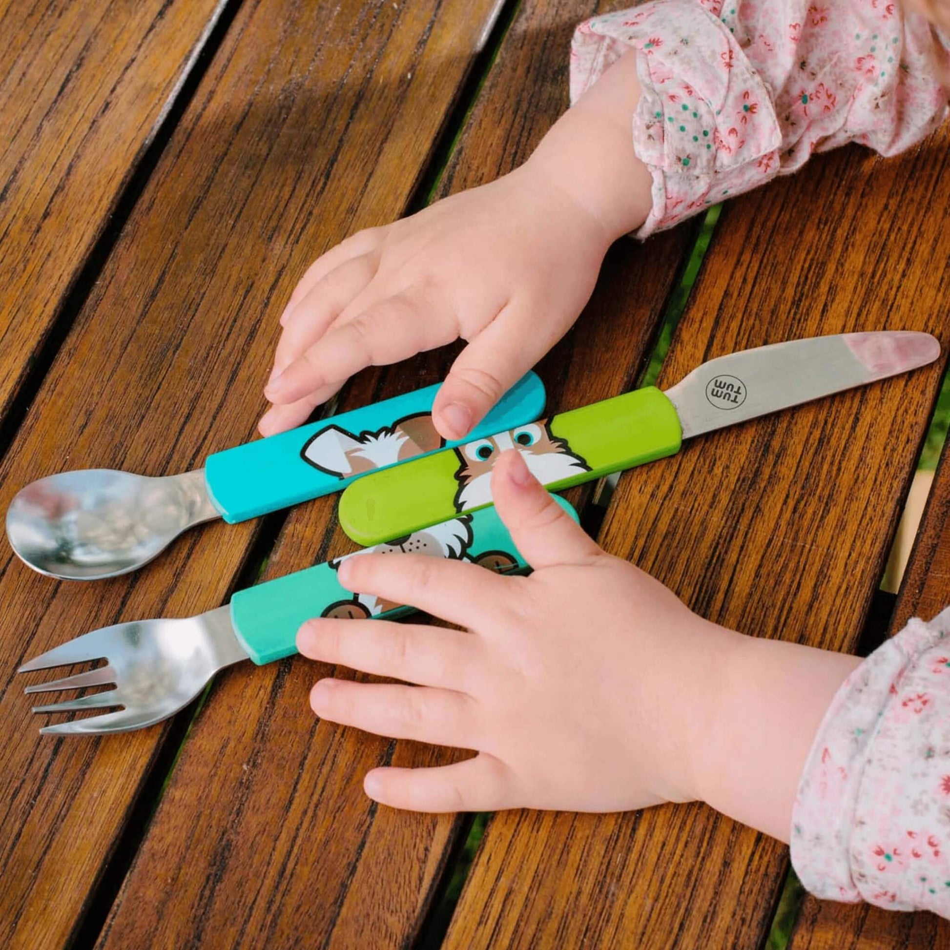 Toddler hands holding a colorful spork and fork set with cartoon animal handles on a wooden table. The utensils have turquoise and green handles with a white dog illustration and a small 'bpb' brand logo on the silver metal blades.