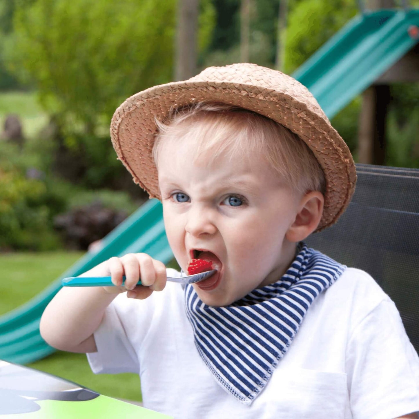 A toddler wearing a straw hat and a blue-and-white striped bib bandana while brushing their teeth outdoors in a garden setting with a green slide in the background.