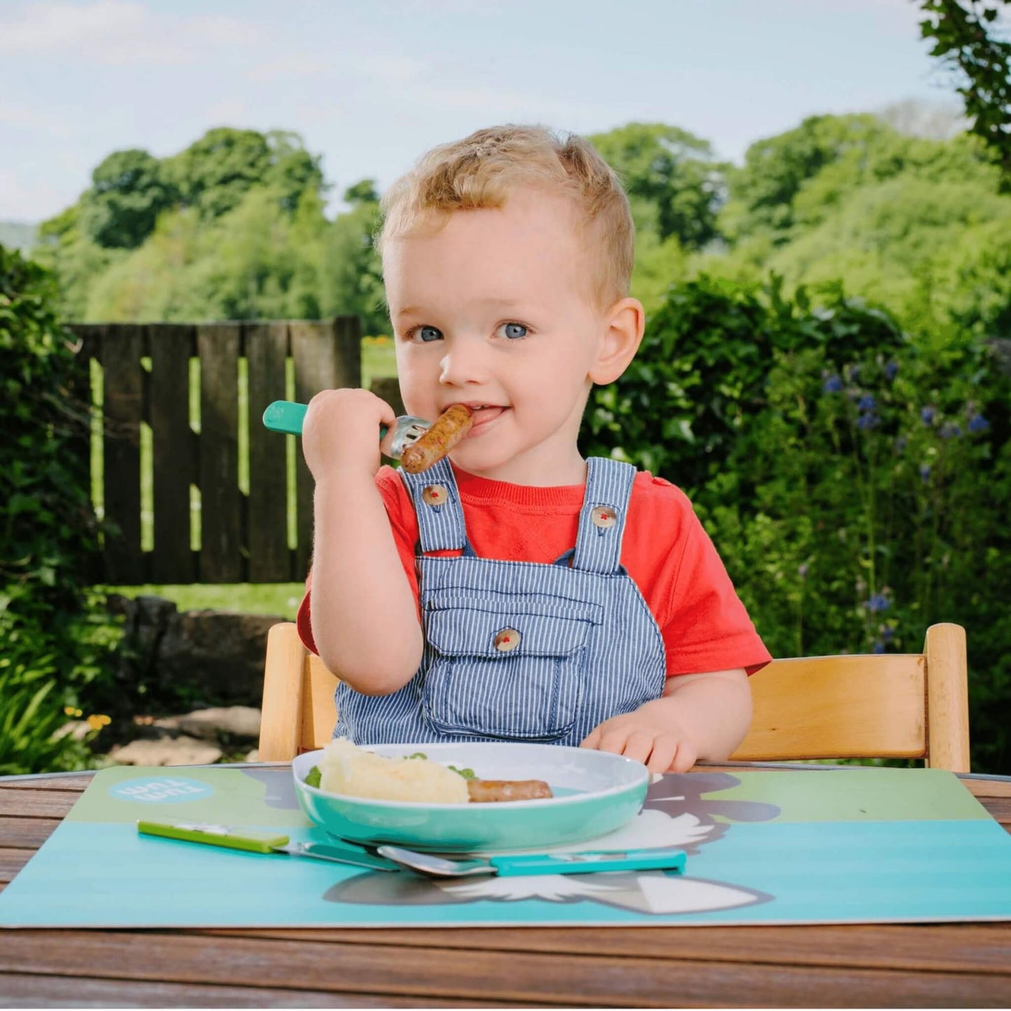A toddler wearing blue denim overalls and a red t-shirt sits at a wooden outdoor table, eating from a light blue bowl. A matching light blue placemat with a cartoon character is on the table, and a green-handled utensil rests near the bowl.