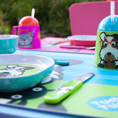 A colorful children's table setting with cartoon dog-themed plates, a green-handled knife, a pink chair, and matching cups and placemats on a blue table outdoors.