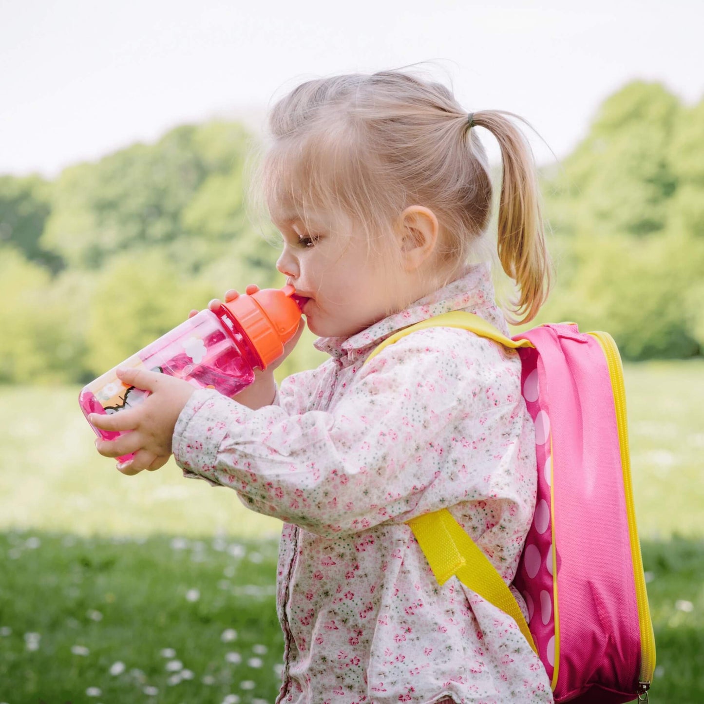A young child wearing a pink and yellow backpack drinks from a pink water bottle outdoors in a grassy park.