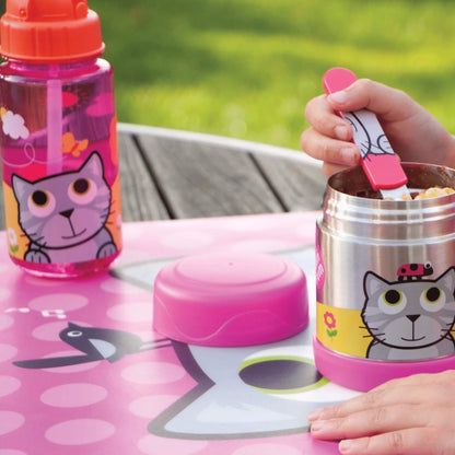 A pink and silver children's lunch set on a pink cat-themed placemat outdoors. The set includes a pink water bottle with a cartoon cat, a stainless steel lunch container with a pink base, a pink lid, and a pink spoon held by a child's hand.