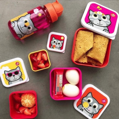 A set of cartoon cat-themed lunch containers and a matching water bottle arranged on a grey surface. The set includes pink, red, yellow, and white containers with crackers, strawberries, an egg, and a snack bar, alongside a pink bottle with an orange cap.