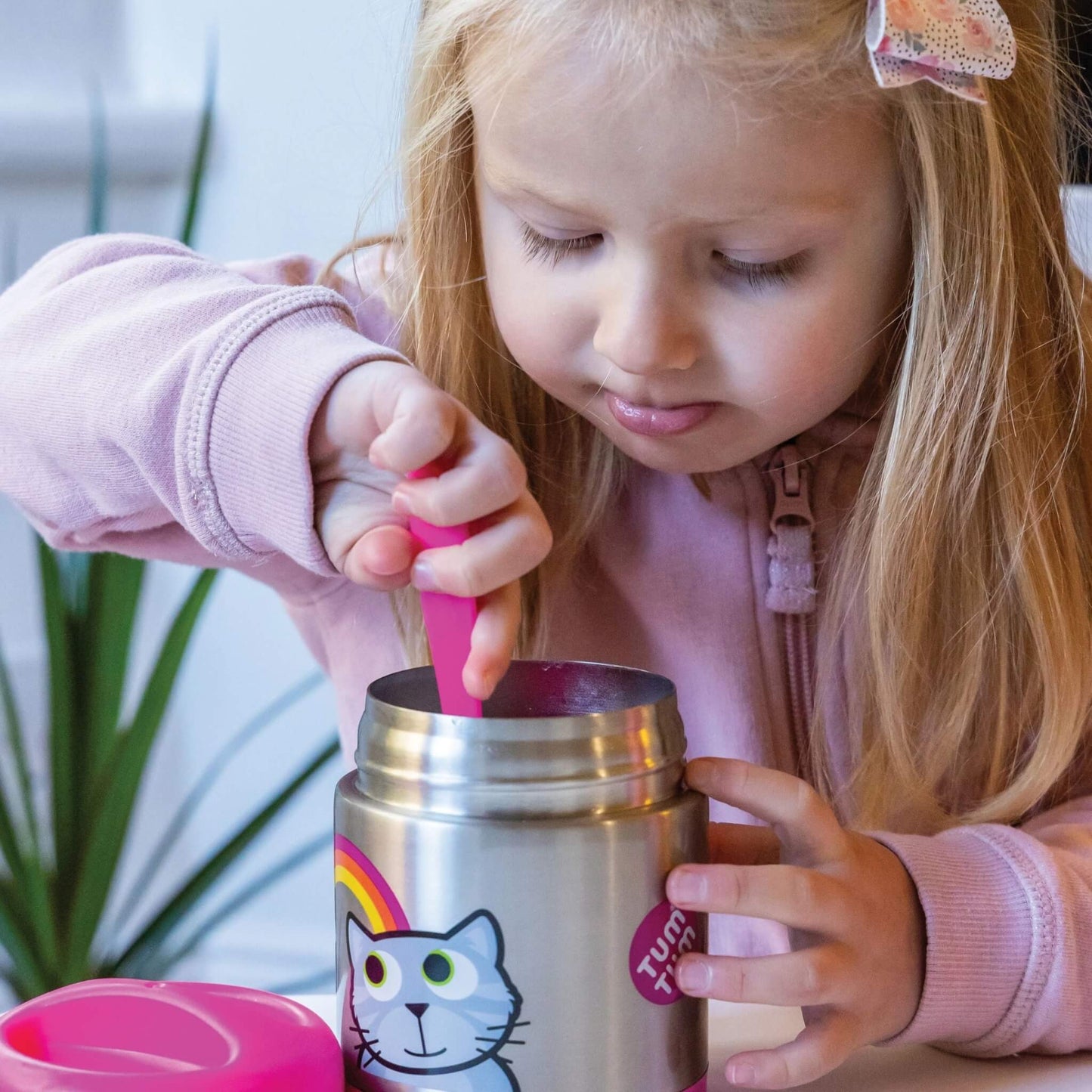 A young girl with blonde hair wearing a pink zip-up hoodie is shown from the side, using a pink spoon to stir a stainless steel tumbler. The tumbler has a cartoon cat face and rainbow graphic on the front, and a pink lid is visible in the foreground.