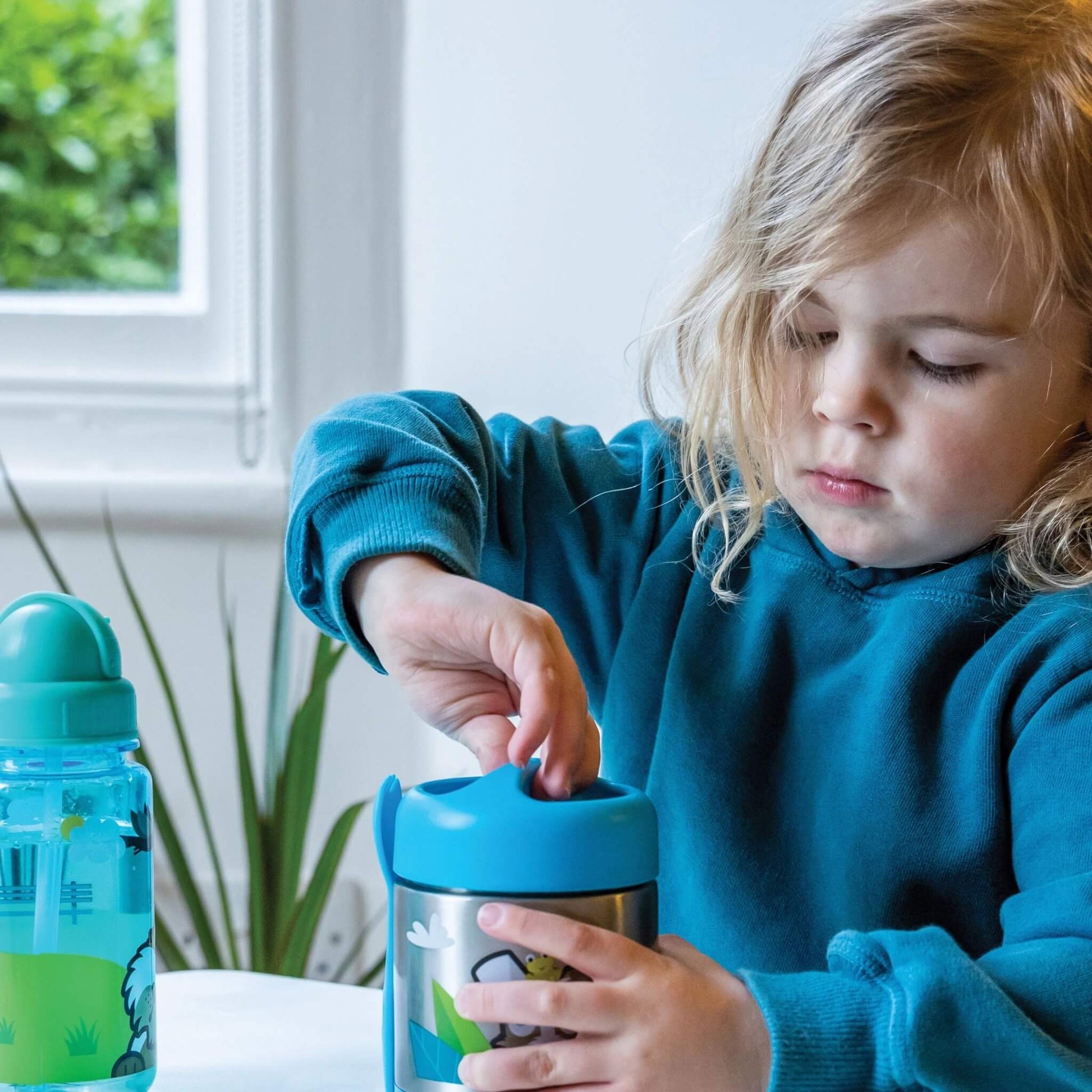 A young child in a teal sweatshirt opens a stainless steel water bottle with a bright blue lid, sitting at a table next to another water bottle.