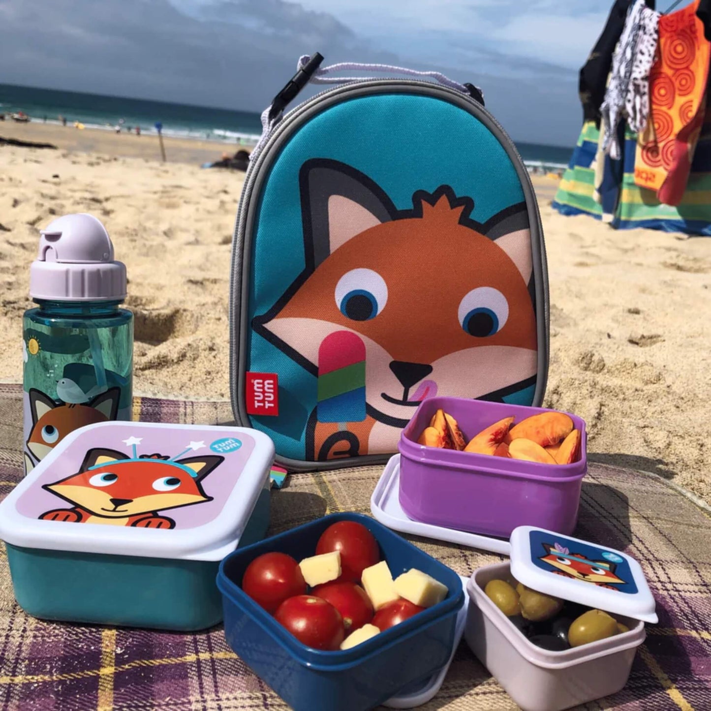 Insulated lunch bag displayed on a picnic blanket with matching food containers and a drinks bottle arranged in front.