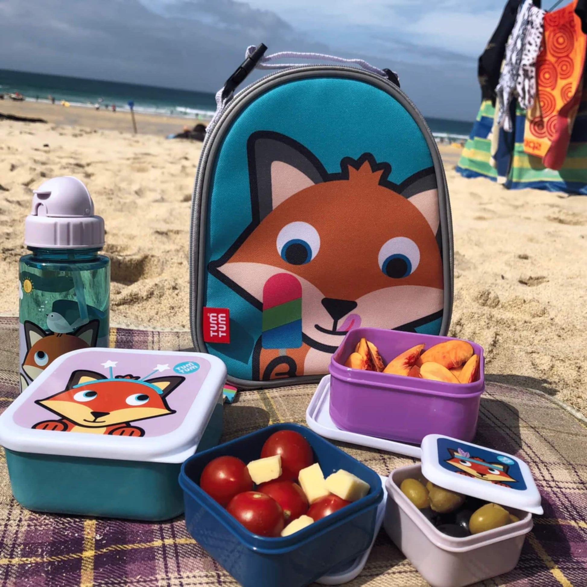 Insulated lunch bag displayed on a picnic blanket with matching food containers and a drinks bottle arranged in front.