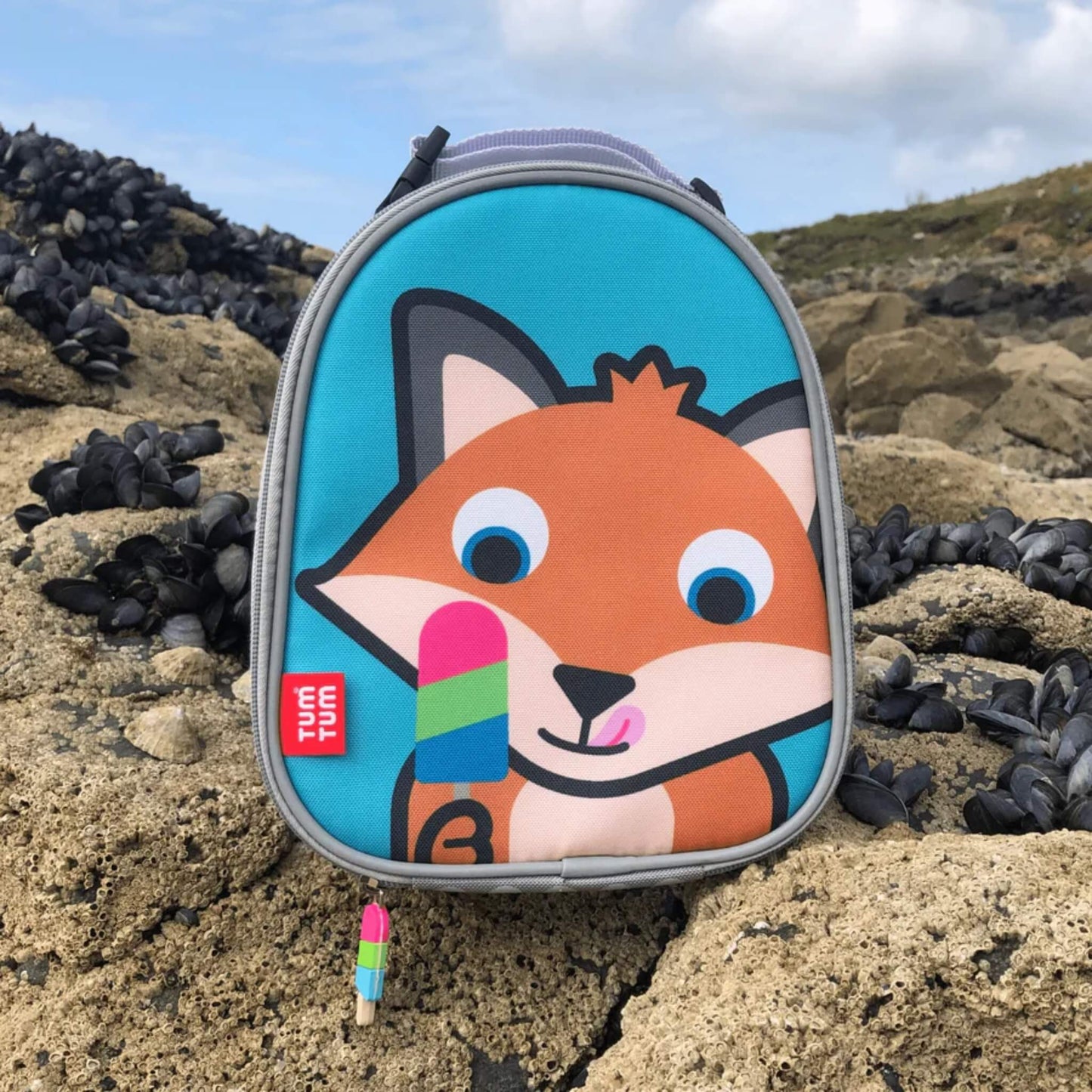Insulated lunch bag standing on rocks outdoors, showing the fox character front panel and zip pull detail at the base.