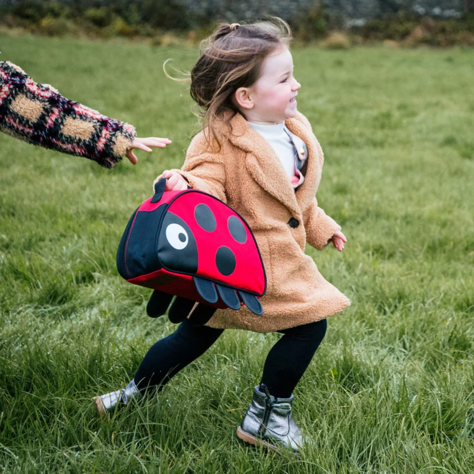 A child running across grass while holding a red ladybird-shaped insulated lunch bag by the carry handle.