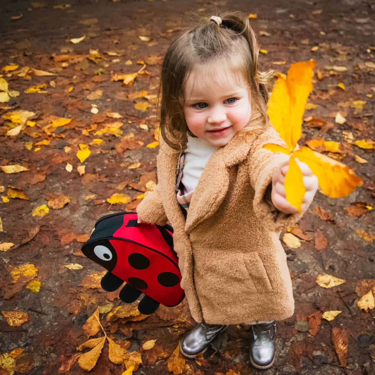 A child standing on a leaf-covered path holding a red ladybird-shaped insulated lunch bag and a yellow leaf, with autumn foliage on the ground.